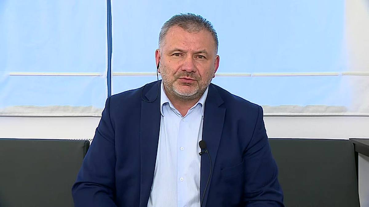 Mężczyzna w średnim wieku w granatowej marynarce i białej koszuli siedzi na kanapie, patrząc w obiektyw kamery.