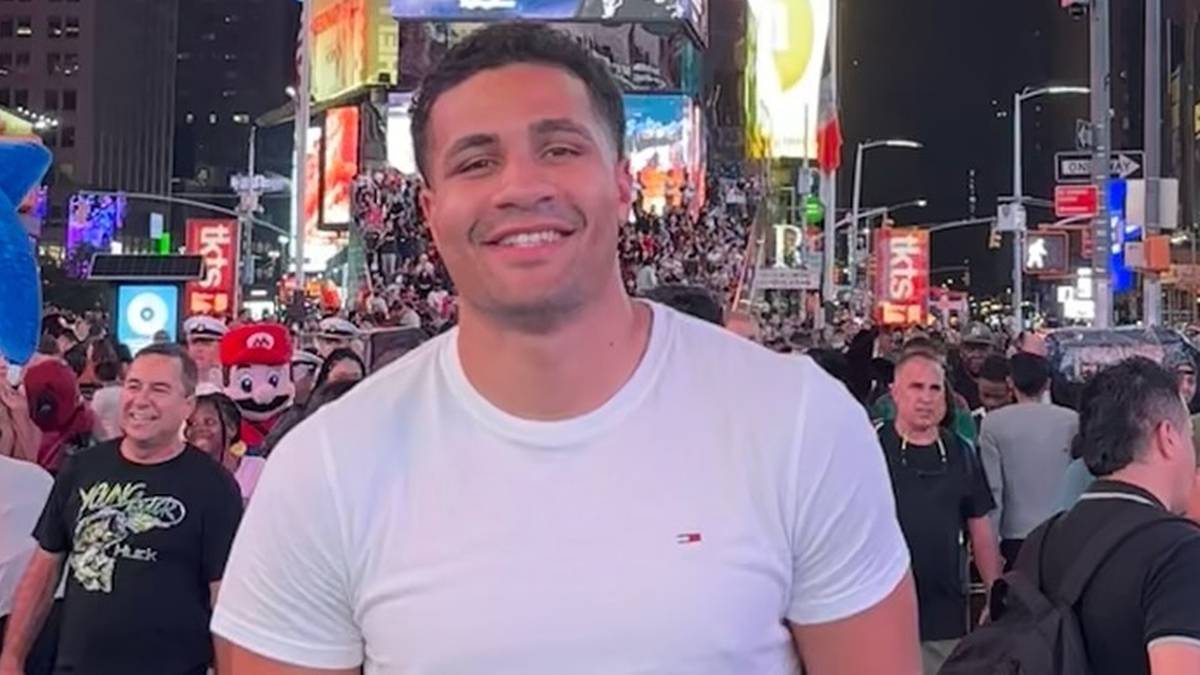 Uśmiechnięty mężczyzna w białej koszulce na tle rozświetlonego wieczornego miasta z widocznymi reklamami i tłumem ludzi.