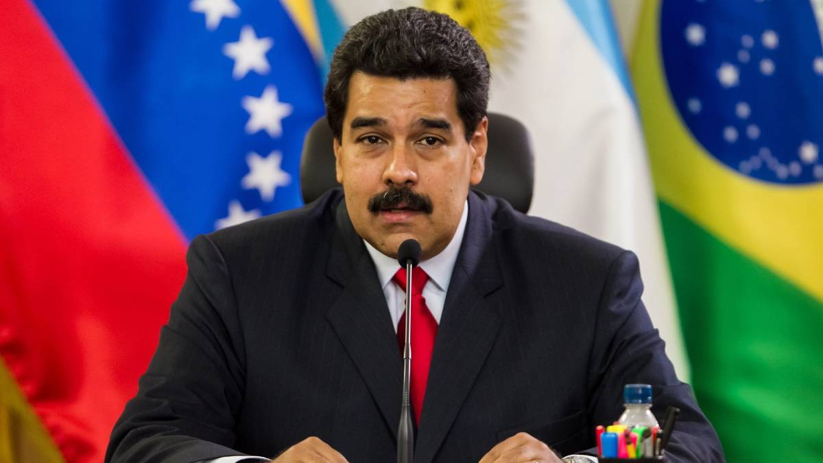 Nicolas Maduro, prezydent Wenezueli, siedzi przy stole z mikrofonem, z flagami Wenezueli, Argentyny i Brazylii w tle.