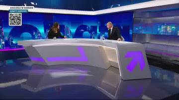 Dwie osoby siedzą przy futurystycznym stole w studio telewizyjnym. Na stole widoczne są fioletowe symbole strzałek. W tle znajduje się niebieskie, nowoczesne tło z elementami graficznymi.