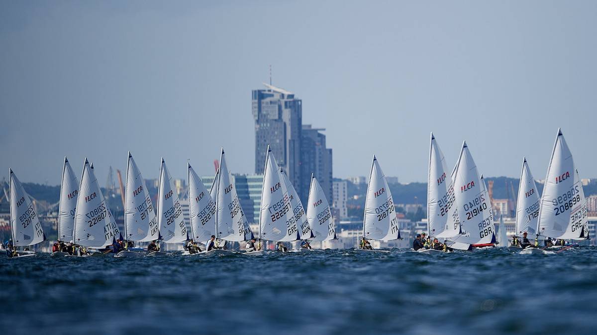 Grupa żagl&oacute;wek ścigających się na wodzie przed miejskim krajobrazem.
