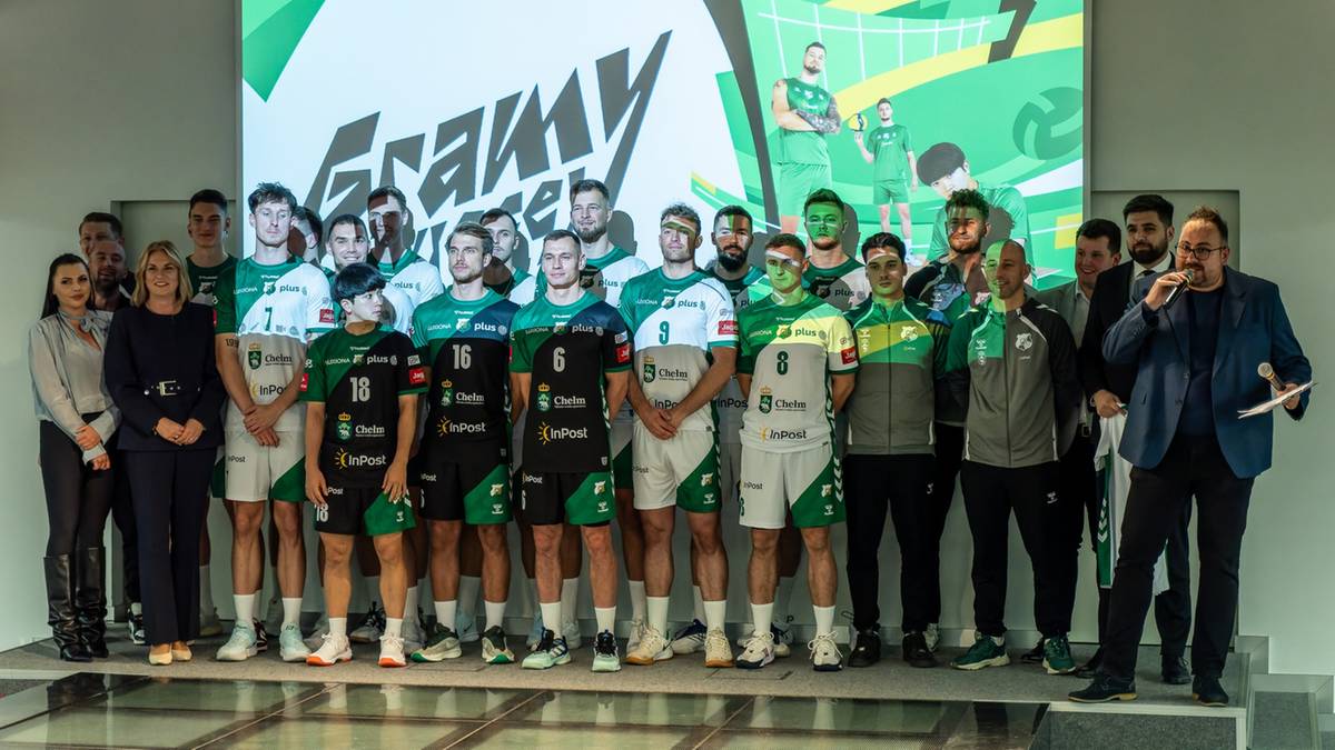 Drużyna siatkarska ChKS Chełm w strojach sportowych pozuje do zdjęcia na tle banera z napisem "Gramy dalej". W tle widoczni są również przedstawiciele klubu.