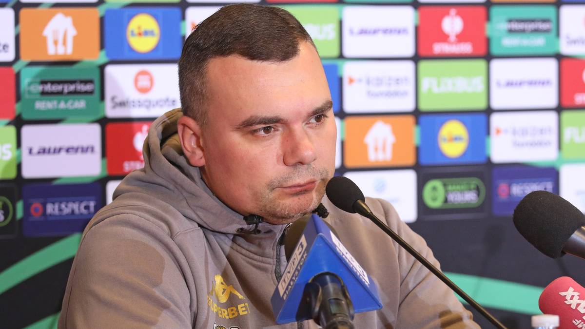 Trener Jagiellonii Adrian Siemieniec siedzi przy stole konferencyjnym, mówiąc do mikrofonu.