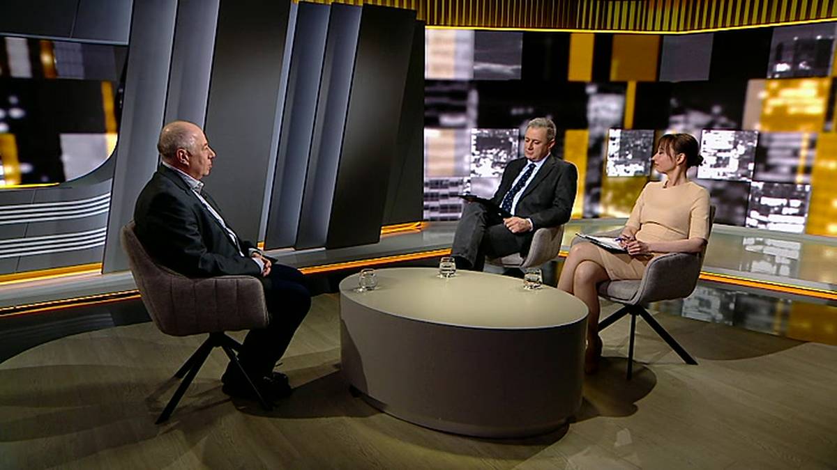 Dw&oacute;ch mężczyzn i kobieta siedzą na krzesłach wok&oacute;ł owalnego stołu w studiu telewizyjnym. Po lewej siedzi starszy mężczyzna w ciemnej marynarce i spodniach. Pośrodku siedzi mężczyzna w garniturze. Po prawej siedzi kobieta w jasnej sukience.