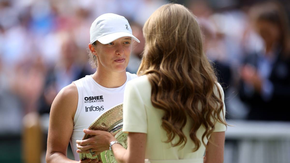 Iga Świątek w białej czapce z daszkiem i białej koszulce z logo OShee i Infosys trzyma trofeum. Rozmawia z kobietą o jasnych włosach.