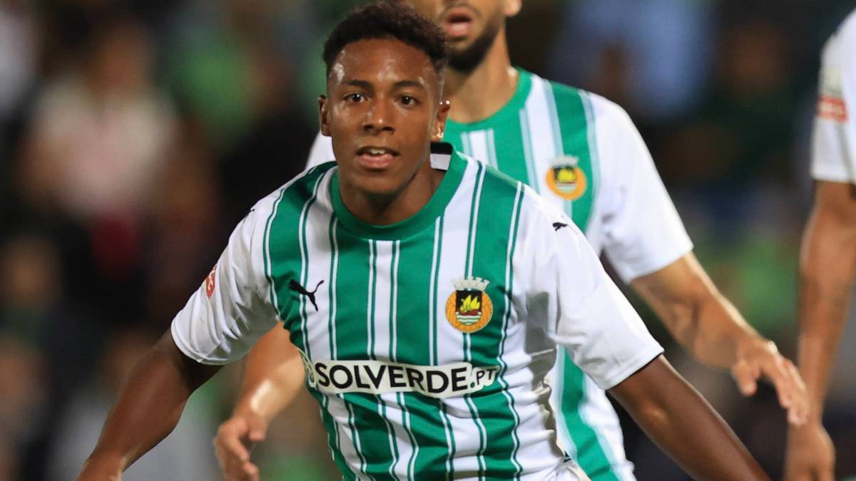 Młody mężczyzna w białej koszulce w zielone pasy z logo klubu piłkarskiego, z napiętą miną patrzy przed siebie.