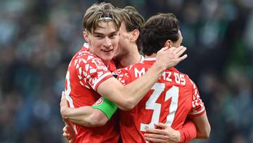 Piłkarz w czerwonej koszulce obejmuje koleg&oacute;w z drużyny, uśmiechając się do kamery.