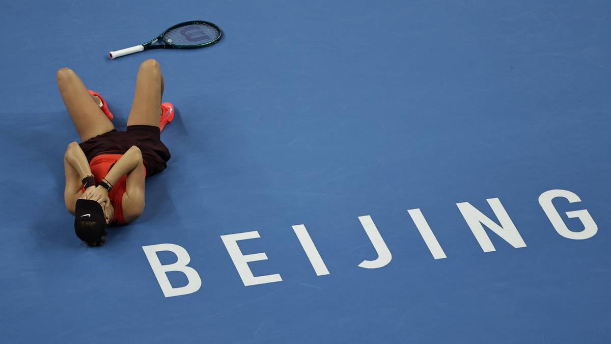 Kobieta w stroju tenisowym leży na niebieskim korcie z rakietą obok, z napisem "BEIJING" w białych literach obok niej.