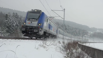 Pociąg PKP Intercity w zimowej scenerii, poruszający się po torach pokrytych śniegiem. W tle widoczne drzewa i wzg&oacute;rza, r&oacute;wnież pokryte śniegiem. Widoczna mgła lub zamieć śnieżna.