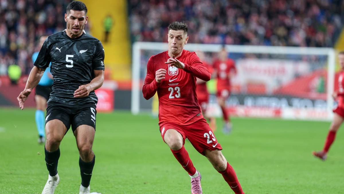 Dwóch piłkarzy podczas meczu piłki nożnej. Piłkarz w czerwonej koszulce z numerem 23 biegnie z piłką, a piłkarz w czarnej koszulce z numerem 5 go kryje. W tle trybuny i bramka.