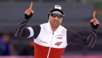 Łyżwiarz szybki w czapce, goglach i kombinezonie z podniesionymi palcami wskazującymi w górę, wyrażający radość.