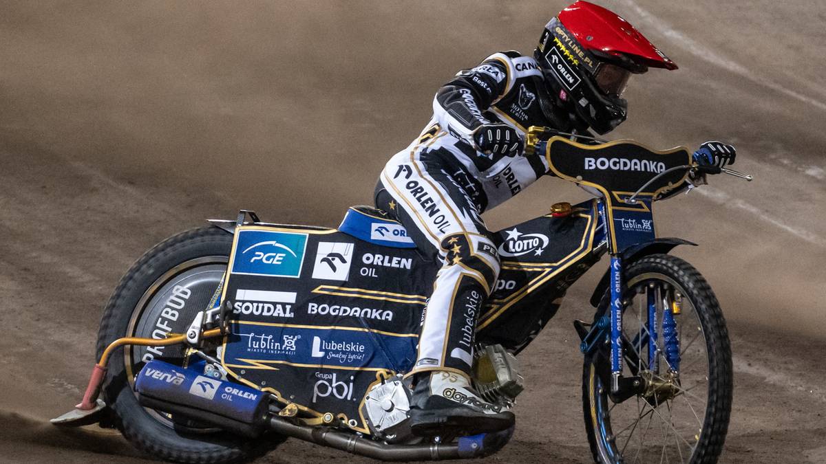 Żużlowiec w kombinezonie i czerwonym kasku ściga się motocyklem po torze.