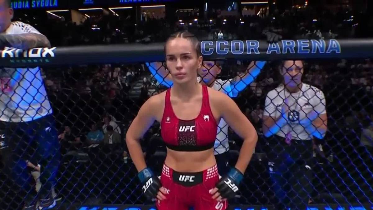 Kobieta w czerwonym stroju sportowym UFC, stojąca na ringu MMA, zdeterminowana.