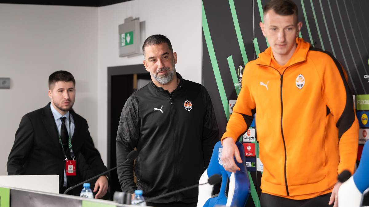 Trzech mężczyzn stoi za stołem konferencyjnym. Dw&oacute;ch z nich to trenerzy w strojach sportowych, trzeci ubrany jest w garnitur.