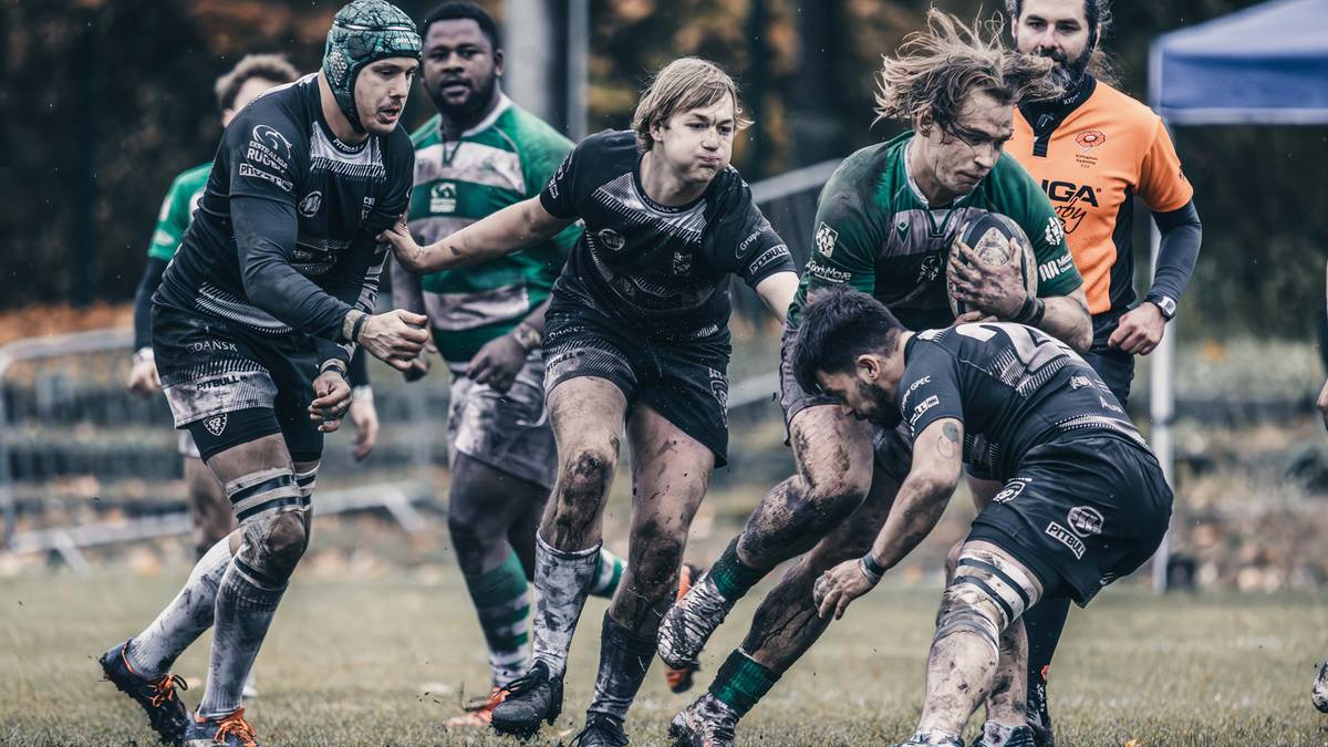 Drużyna graczy rugby w zielono-czarnych strojach walczy o piłkę na boisku. Jeden z graczy w zielonym stroju próbuje odebrać piłkę przeciwnikowi.