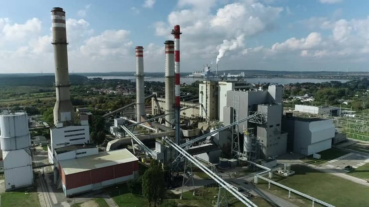 Widok z lotu ptaka na kompleks elektrowni z wieloma kominami, z kt&oacute;rych jeden emituje dym. Widoczne budynki przemysłowe i sieć rurociąg&oacute;w.