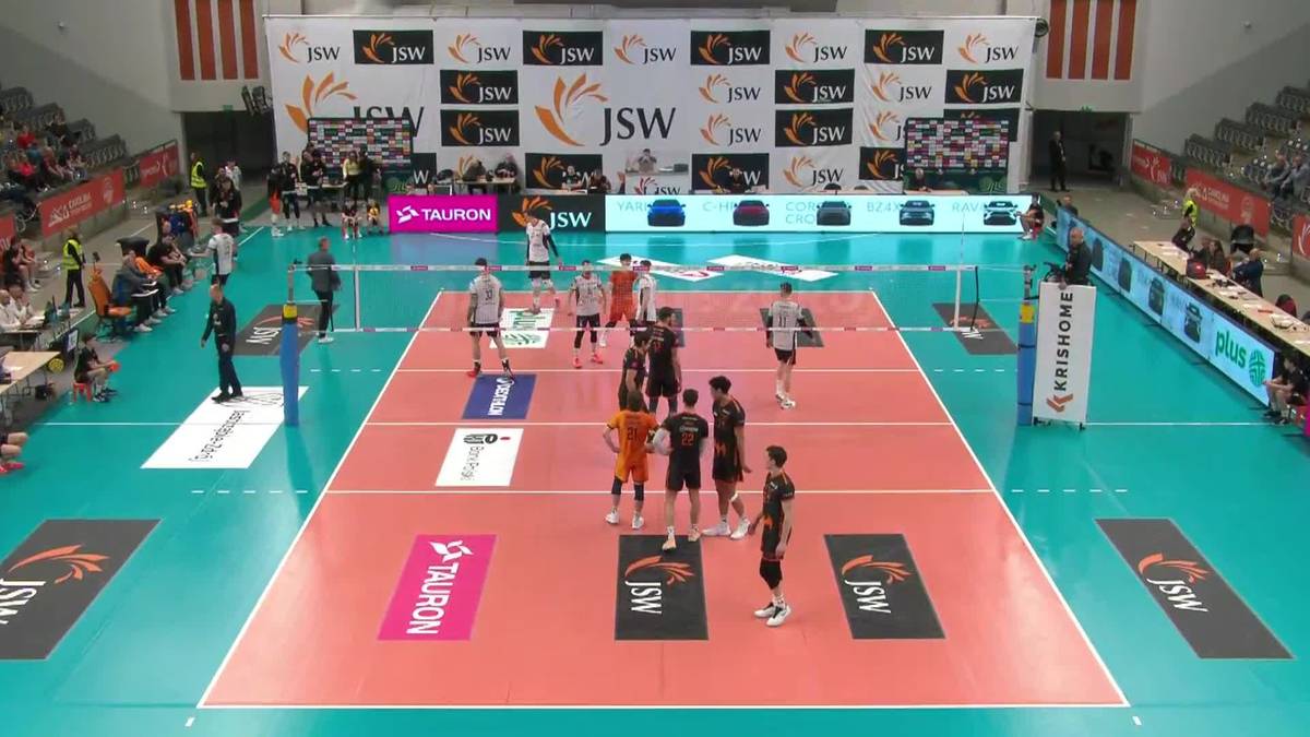 Drużyny siatkarzy w czerwonych i pomarańczowych strojach ustawione na boisku hali sportowej, gotowe do gry, z widownią na trybunach w tle.