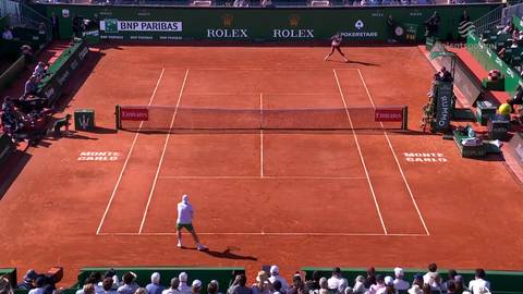 Gracz w białej koszulce i zielonych spodenkach przygotowuje się do uderzenia piłki na korcie ziemnym. W tle widać drugiego gracza, siatkę tenisową i trybuny z widzami.