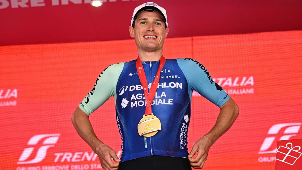 Kolarz z medalem na szyi stoi na podium, nosząc strój kolarski i czapkę.