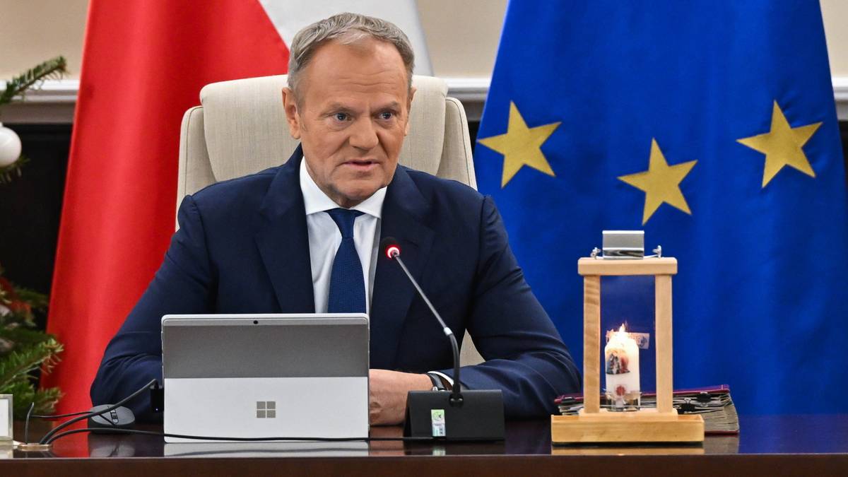Donald Tusk siedzi przy stole, przed nim tablet i mikrofon, w tle flagi Polski i Unii Europejskiej.
