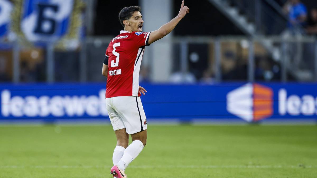 Piłkarz w czerwonej koszulce z numerem 5 i białych spodenkach, stojący na boisku piłkarskim, unoszący prawą rękę z wyprostowanym palcem wskazującym.