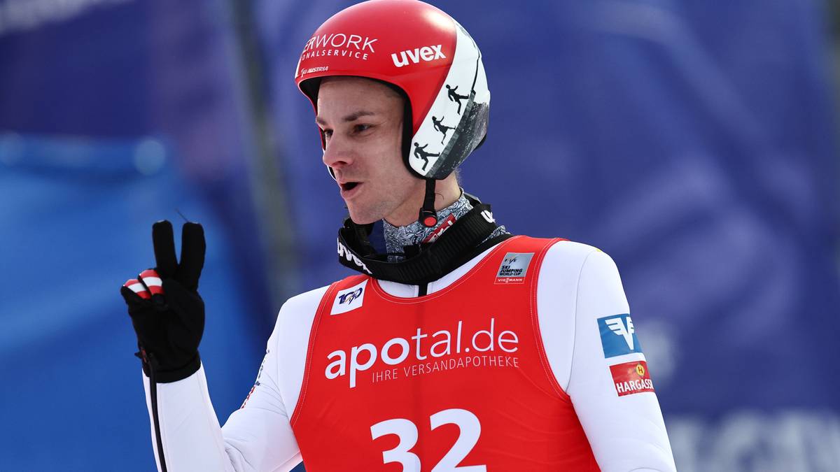 Skoczek narciarski w czerwonym kasku i białej kamizelce sportowej z numerem 32, pokazuje dwa palce w geście zwycięstwa lub pokoju.