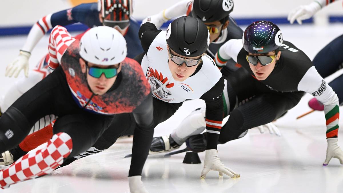 Grupa łyżwiarzy szybkich w pozycjach startowych na lodzie, w kolorowych strojach i kaskach.