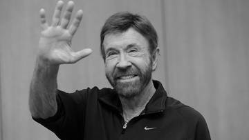 Chuck Norris uśmiecha się i macha prawą ręką.