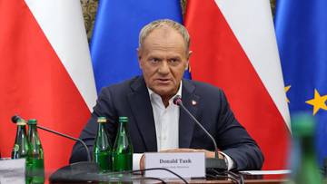 Szef rządu Donald Tusk siedzi przy stole konferencyjnym na tle flag Polski i Unii Europejskiej podczas narady.