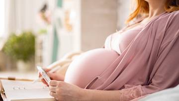 Kobieta w ciąży siedzi, trzymając w dłoniach mały przedmiot, na pierwszym planie widoczny zaokrąglony brzuch.