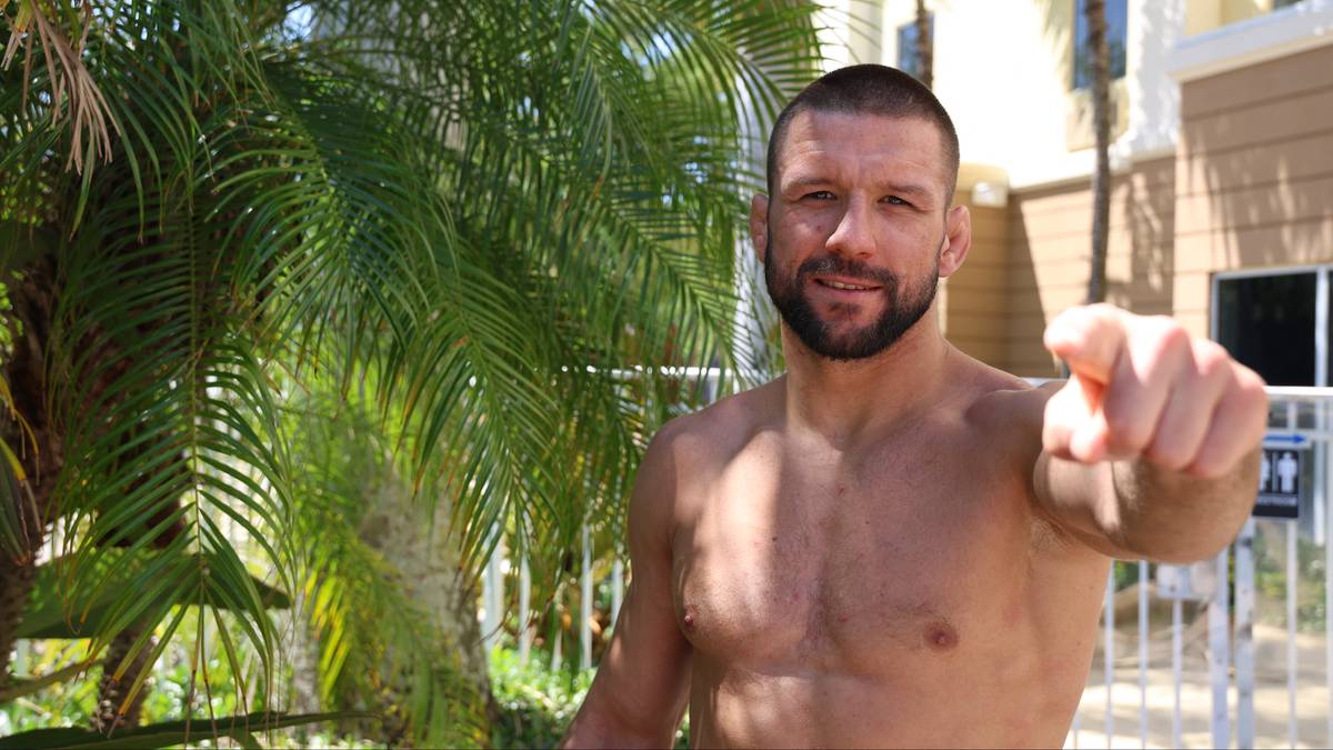 Mężczyzna z krótką brodą, bez koszulki, wskazuje palcem w stronę widza. W tle widoczne palmy i budynek.