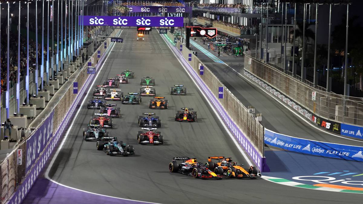 Widok z g&oacute;ry na grupę samochod&oacute;w Formuły 1 ścigających się na torze nocą, z jaskrawymi światłami otaczającymi trasę.