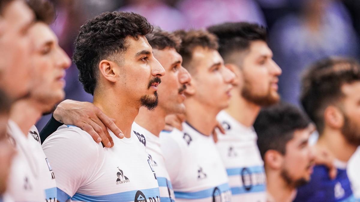 Portret grupowy siatkarzy w białych koszulkach z niebieskimi elementami, skupiony na profilu zawodnika z ciemnymi włosami i wąsami.