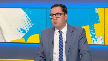 Mężczyzna w niebieskim garniturze i okularach siedzi w studiu telewizyjnym.