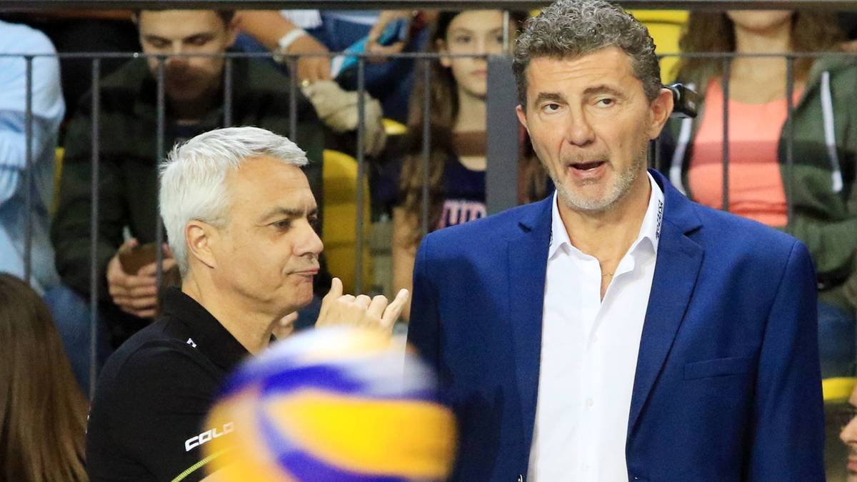 Dwóch mężczyzn w strojach sportowych stoi obok siebie, jeden z nich trzyma tablicę z notatkami, drugi patrzy w kierunku piłki do siatkówki.