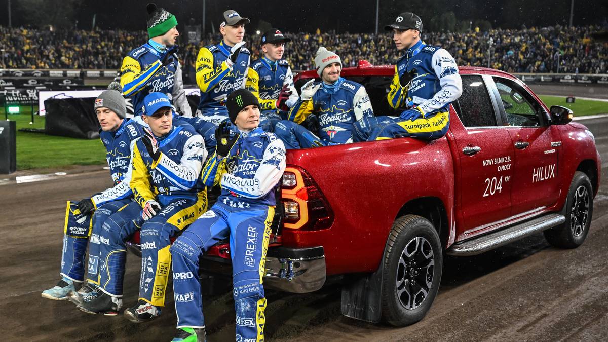 Drużyna żużlowców w niebiesko-żółtych kombinezonach siedzi na pace czerwonego pickupa, pozując do zdjęcia po zwycięstwie.