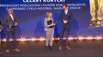 Na scenie stoją trzy osoby podczas ceremonii wręczenia nagród. Po lewej mężczyzna, w środku kobieta, a po prawej mężczyzna z nagrodą. W tle znajduje się napis "CEZARY KORYCKI" i "PRODUCENT PODCASTÓW I FILMÓW DOKUMENTALNYCH, GOSPODARZ CYKLU HISTORIA, JAKIEJ NIE ZNACIE". Scena jest ozdobiona migoczącymi światłami.