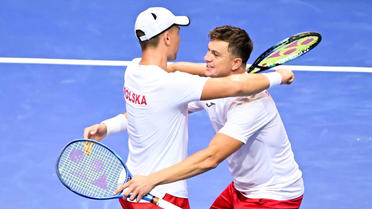 Dwóch tenisistów w białych koszulkach z czerwonym napisem "POLSKA" na plecach, uśmiechają się i przytulają na korcie tenisowym, trzymając rakiety.