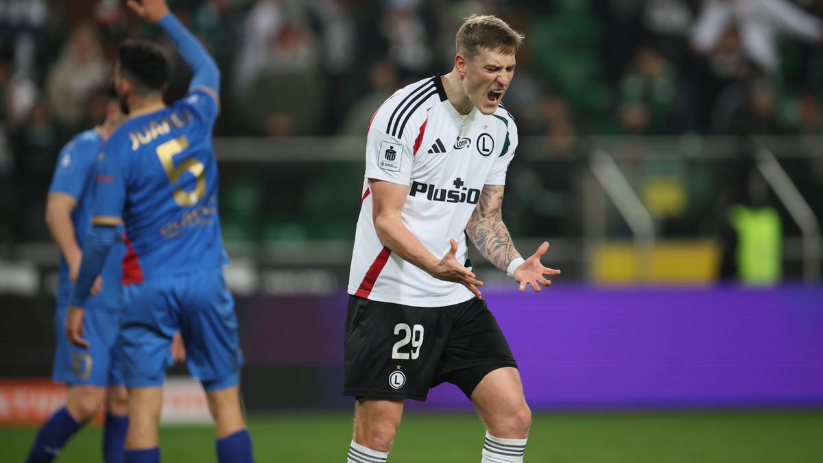 Piłkarz Legii Warszawa w białej koszulce z numerem 29 na plecach, z wykrzywioną twarzą i rozłożonymi rękami, wyrażający frustrację.