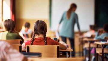 Klasa szkolna z uczniami siedzącymi w ławkach, w tle nauczycielka przy tablicy.