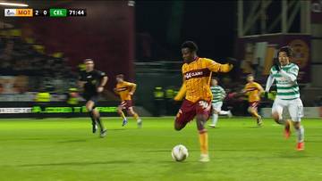 Zawodnicy drużyny Motherwell FC i Celtic FC podczas meczu piłki nożnej. Jeden z graczy Motherwell FC, ubrany w żółto-bordowy strój, prowadzi piłkę, podczas gdy zawodnik Celtic FC w zielono-białym stroju próbuje go zatrzymać.