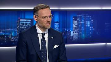 Mężczyzna w garniturze i okularach siedzi i m&oacute;wi, na tle ekran&oacute;w z widokiem nocnego miasta.