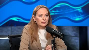 Kobieta w beżowym żakiecie siedzi przy stole z mikrofonem przed sobą.