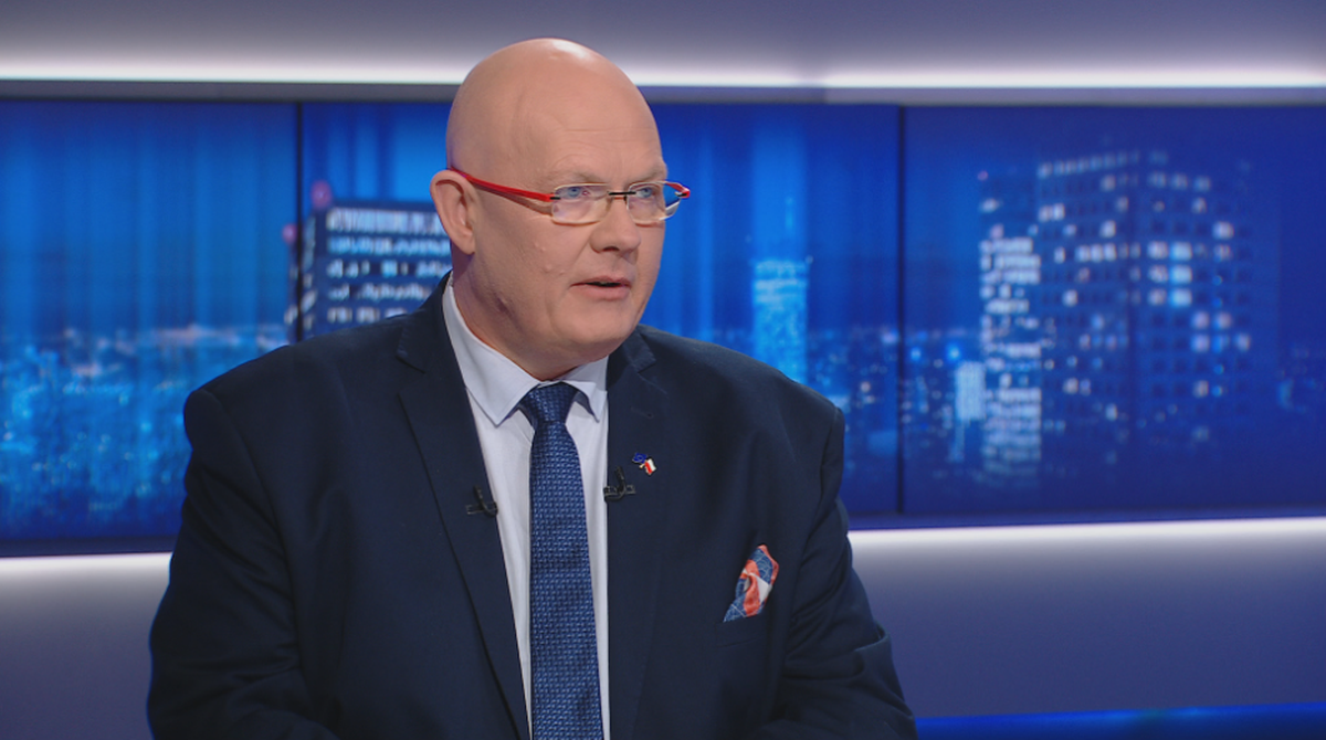 Mężczyzna w okularach i garniturze siedzi przed ekranami.