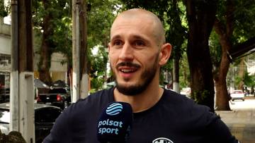 Mężczyzna z ogoloną głową i zarostem, ubrany w ciemną koszulkę, trzyma mikrofon z logo Polsat Sport.