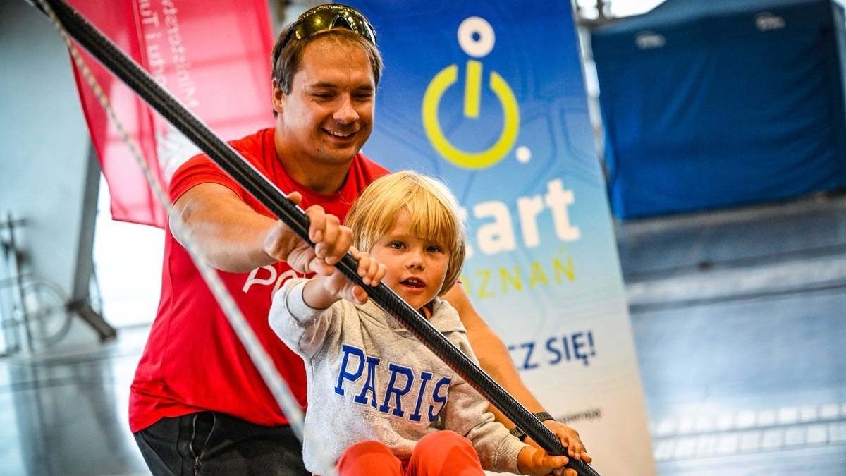 Mężczyzna w czerwonym t-shircie pomaga dziecku trzymać drążek podczas ćwiczeń.