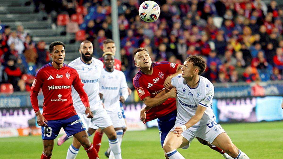 Piłkarze w białych i czerwonych strojach walczą o piłkę na zielonej murawie boiska piłkarskiego. W tle widzowie na trybunach.