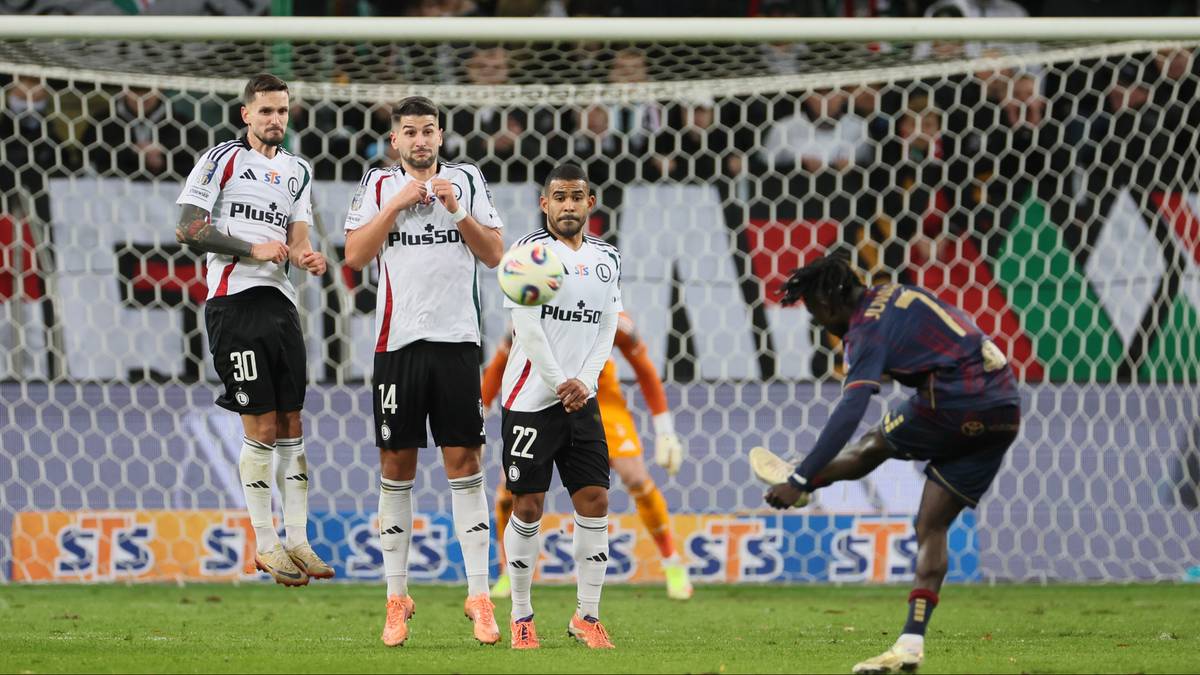 Piłkarze w białych strojach stoją w murze przed bramką, a jeden zawodnik w ciemnym stroju wykonuje rzut karny.