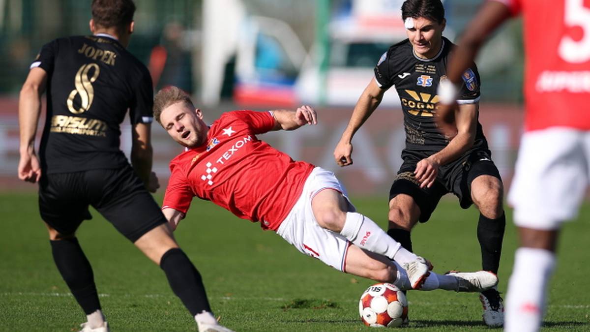 Piłkarz w czerwonej koszulce drybluje z piłką, podczas gdy dwaj zawodnicy w czarnych strojach pr&oacute;bują odebrać mu futbol&oacute;wkę.