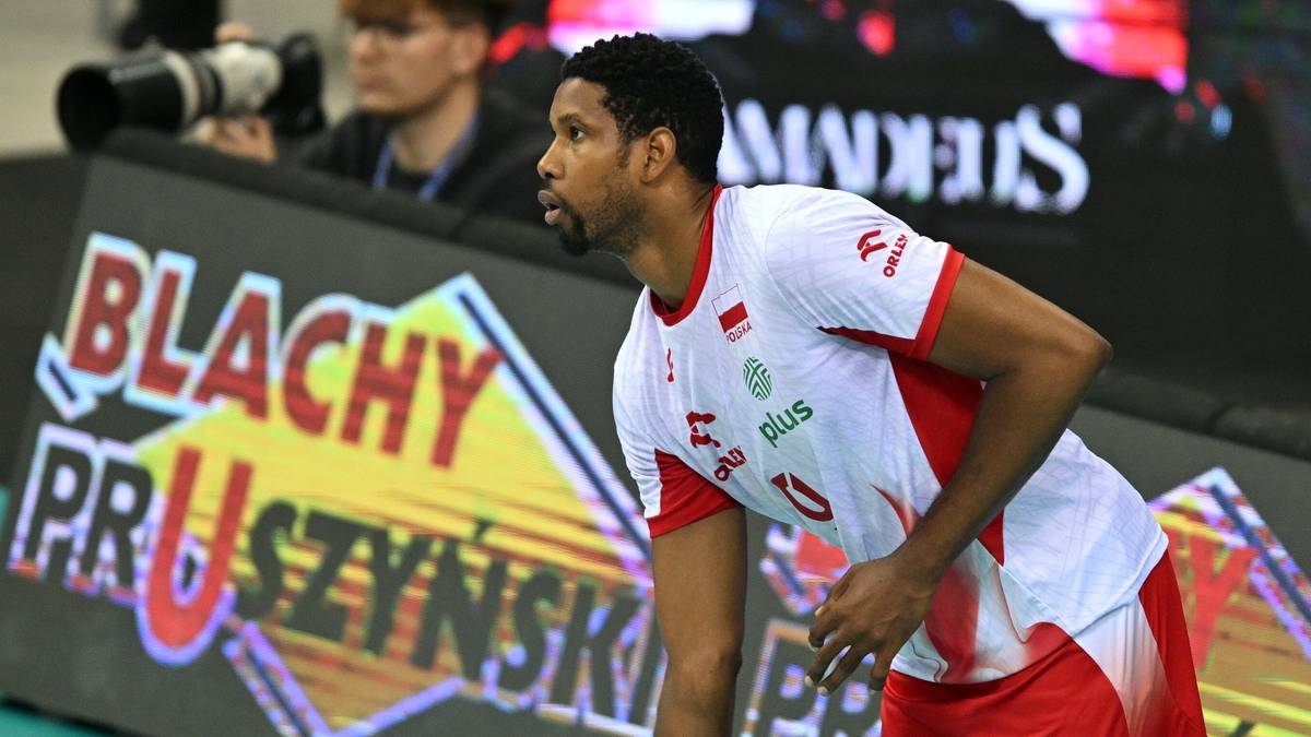 Siatkarz w białej koszulce z czerwonymi wstawkami i numerem 10, na tle band reklamowych.